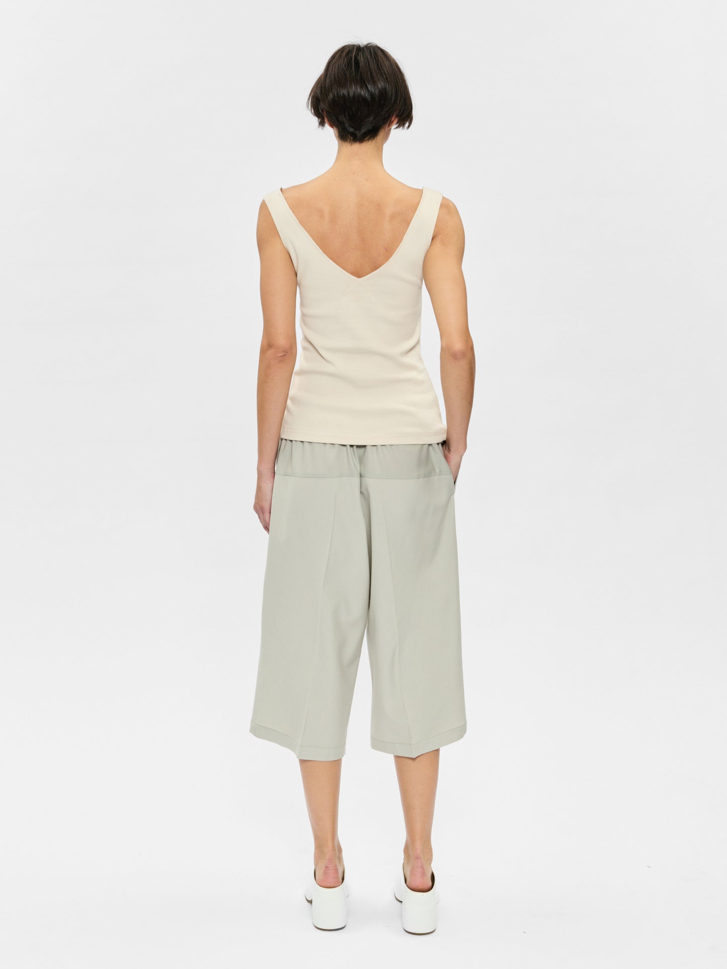 Woman wearing a white sleeveless top and light green pants on a plain background