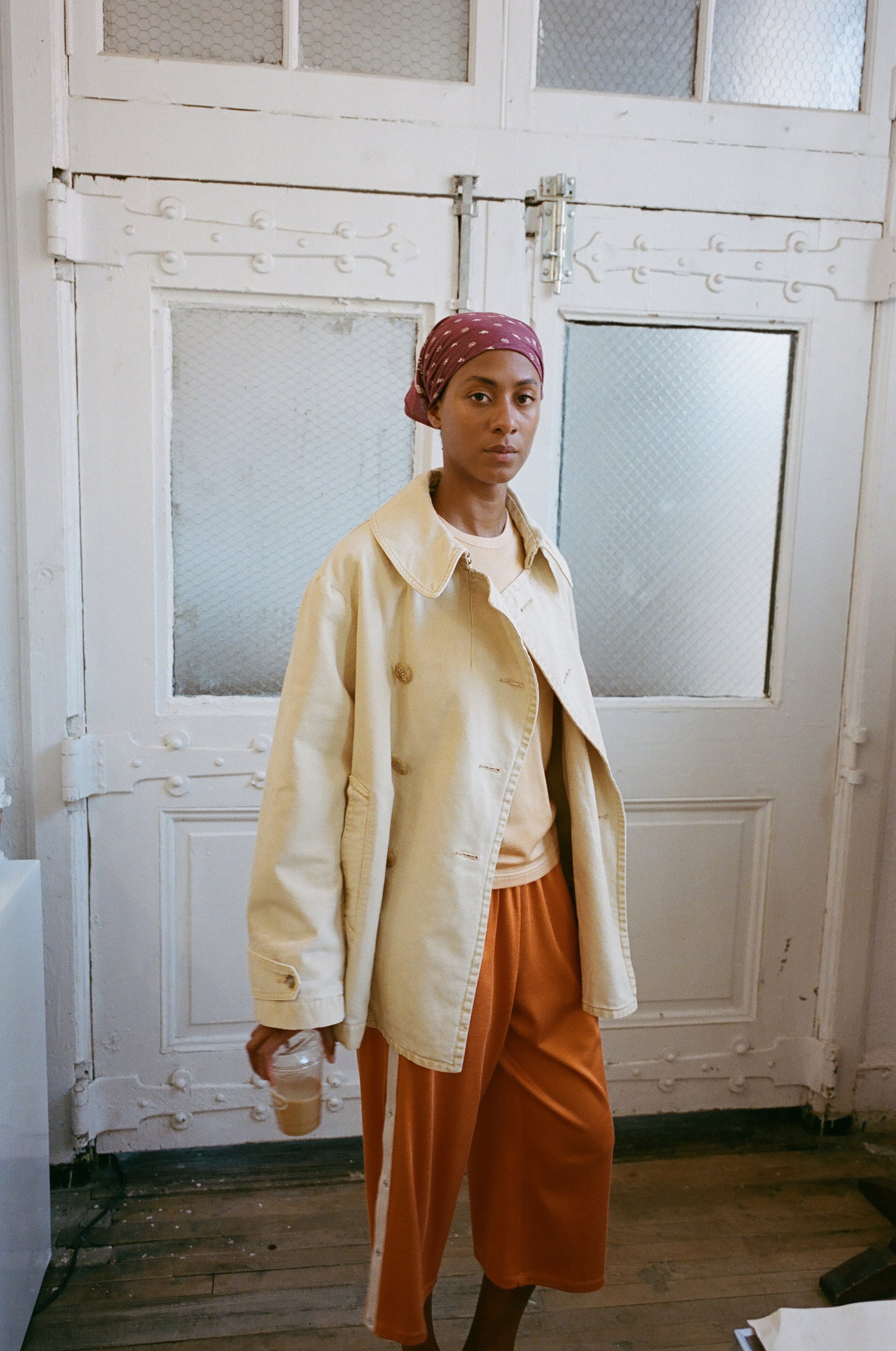 Person wearing a beige jacket and orange pants standing in a room with white walls and a door.