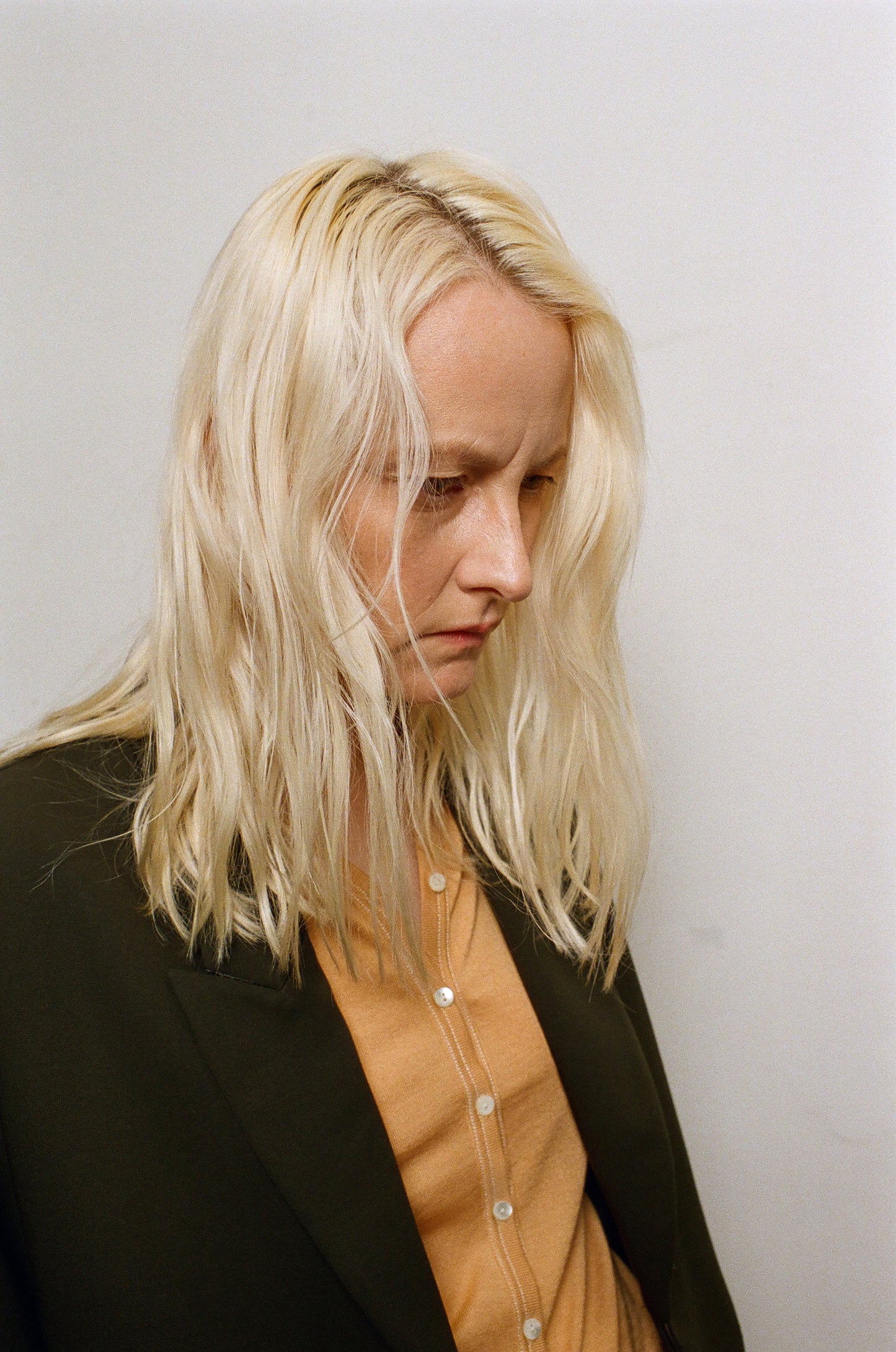 Person with blonde hair wearing a black jacket and beige shirt against a plain background