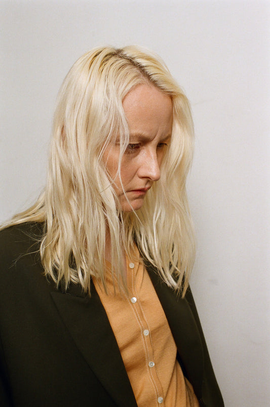 Person with blonde hair wearing a black jacket and beige shirt against a plain background