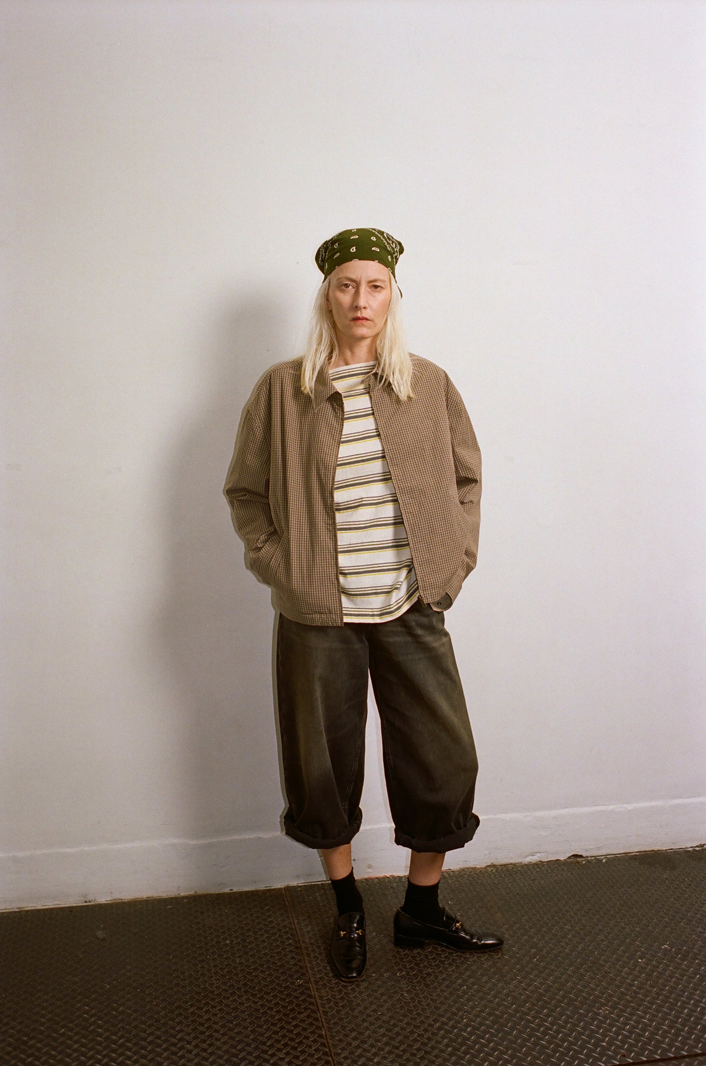 Person wearing a beige jacket, striped shirt, and green pants against a white wall.