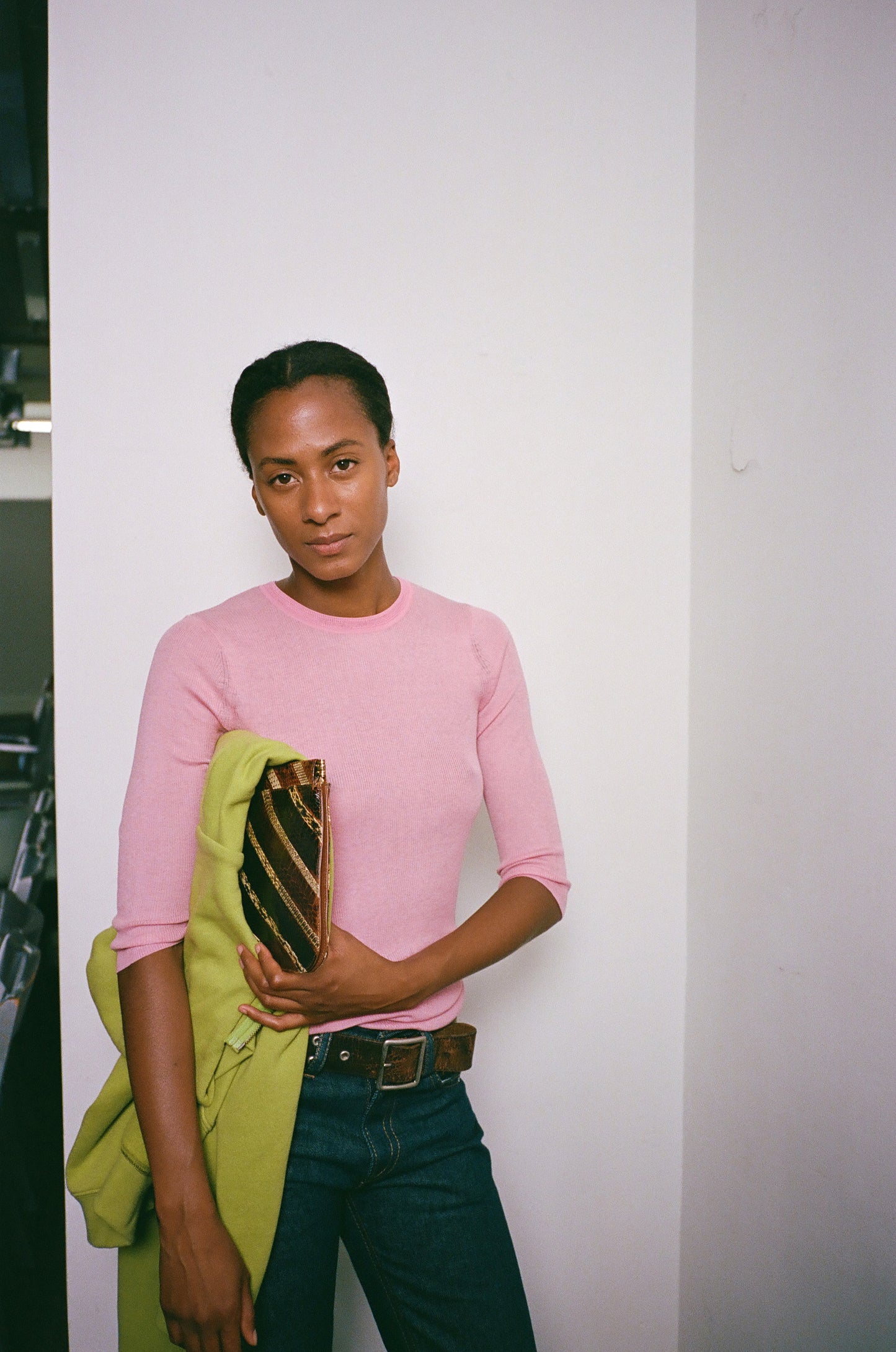 Person wearing a pink sweater and holding a green jacket against a white wall.