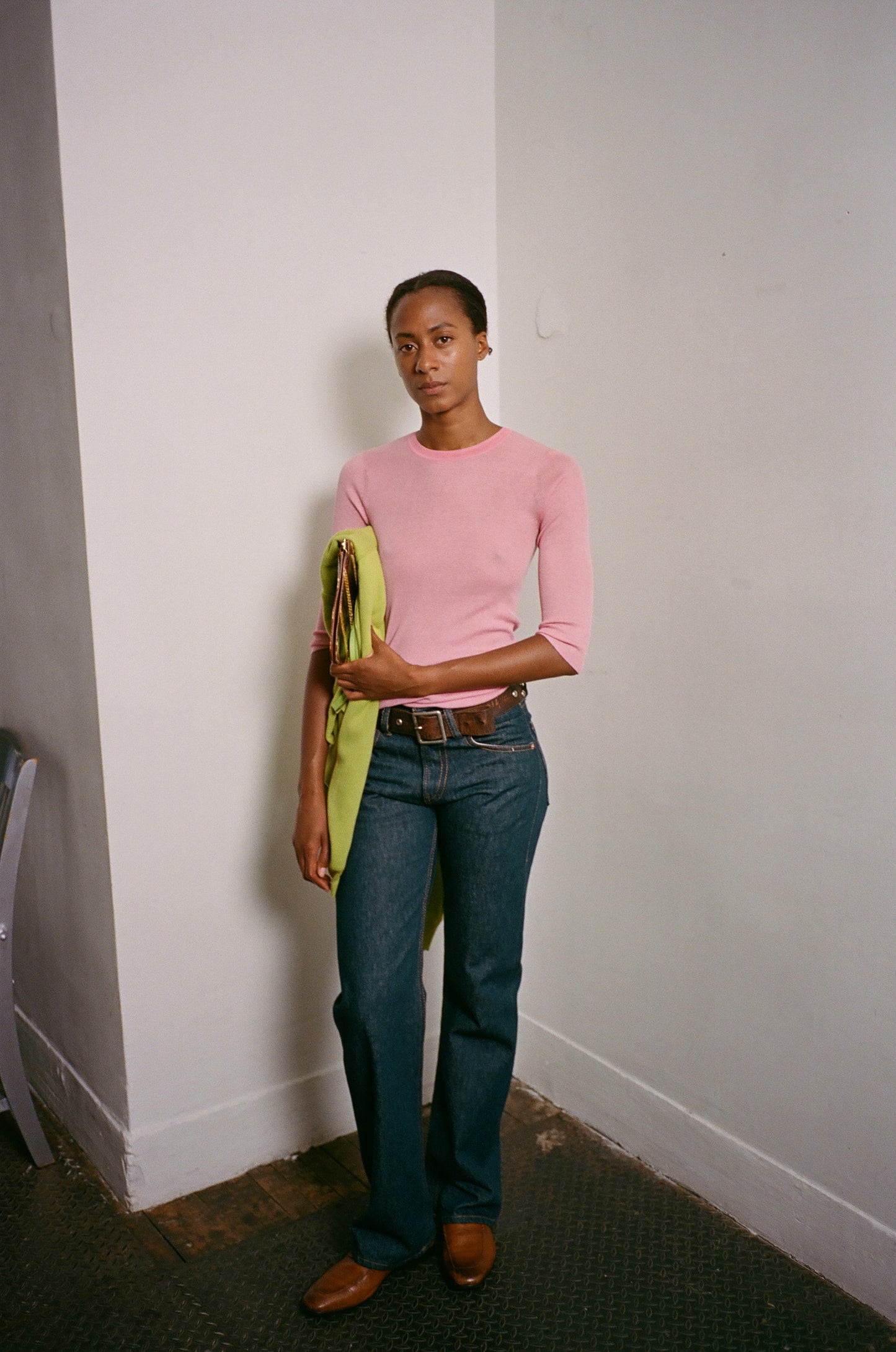 Person wearing a pink sweater and blue jeans holding a green jacket against a white wall.