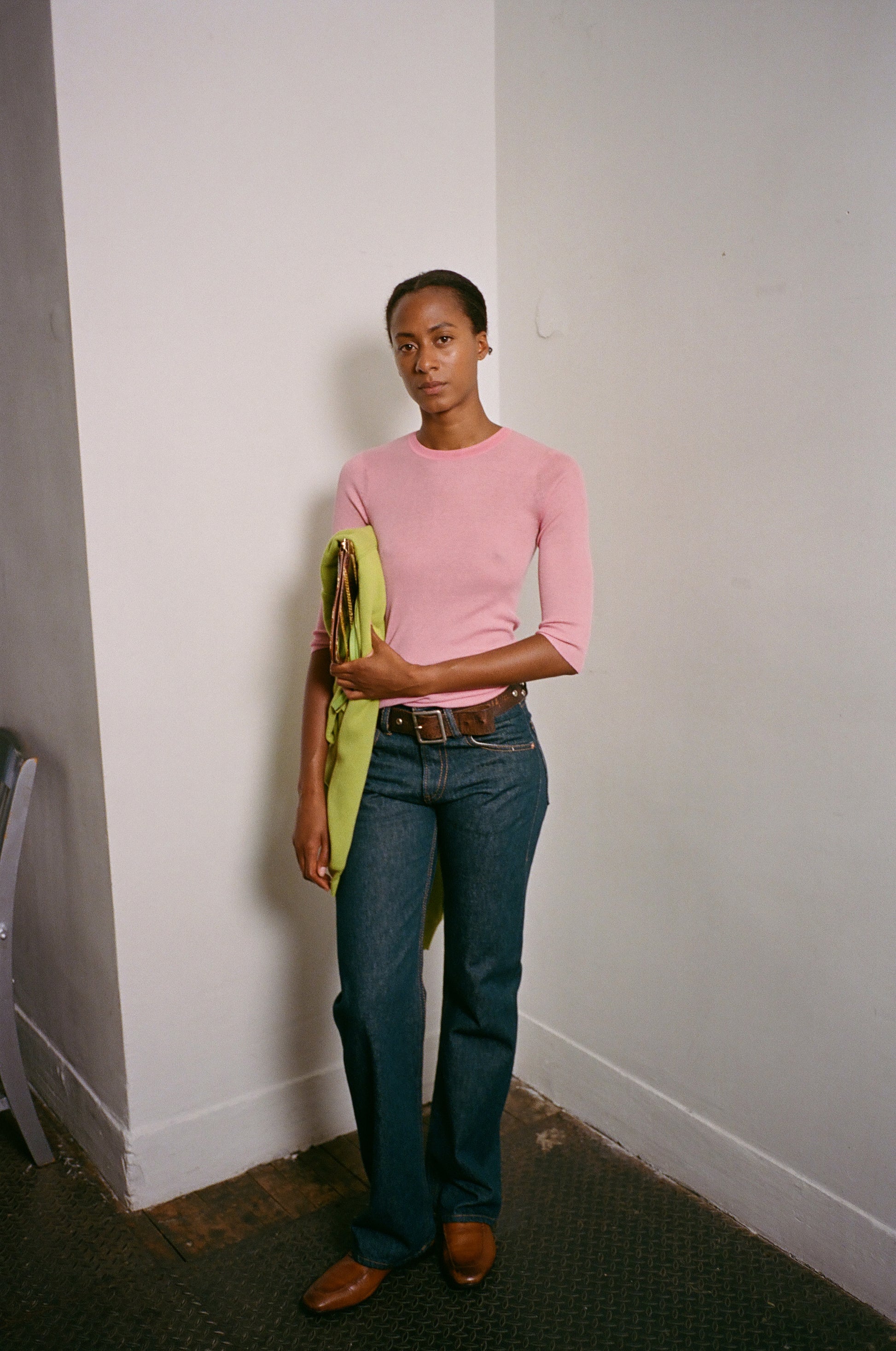Person wearing a pink sweater and blue jeans holding a green jacket against a white wall.