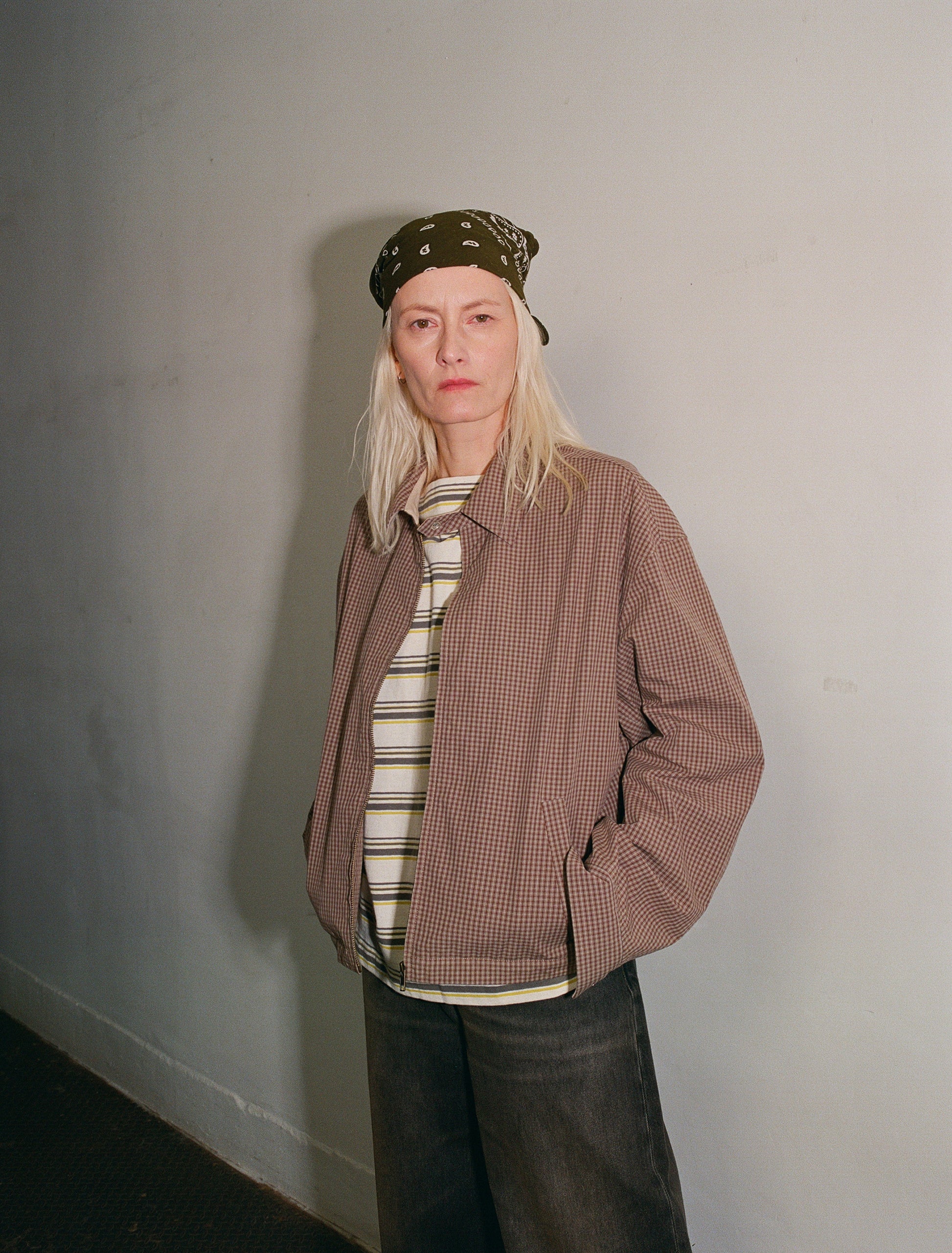 Person wearing a brown checkered jacket, striped shirt, and dark pants against a plain wall.