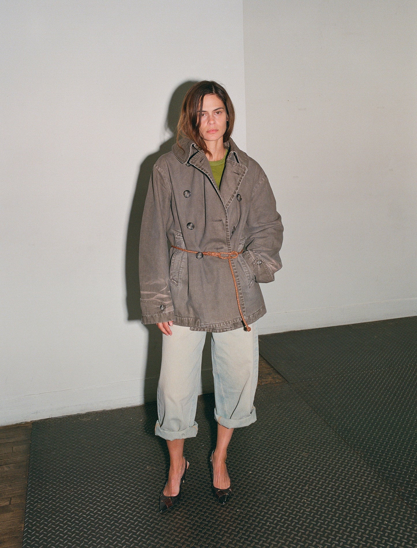 Person wearing a brown coat and light blue jeans standing against a white wall.