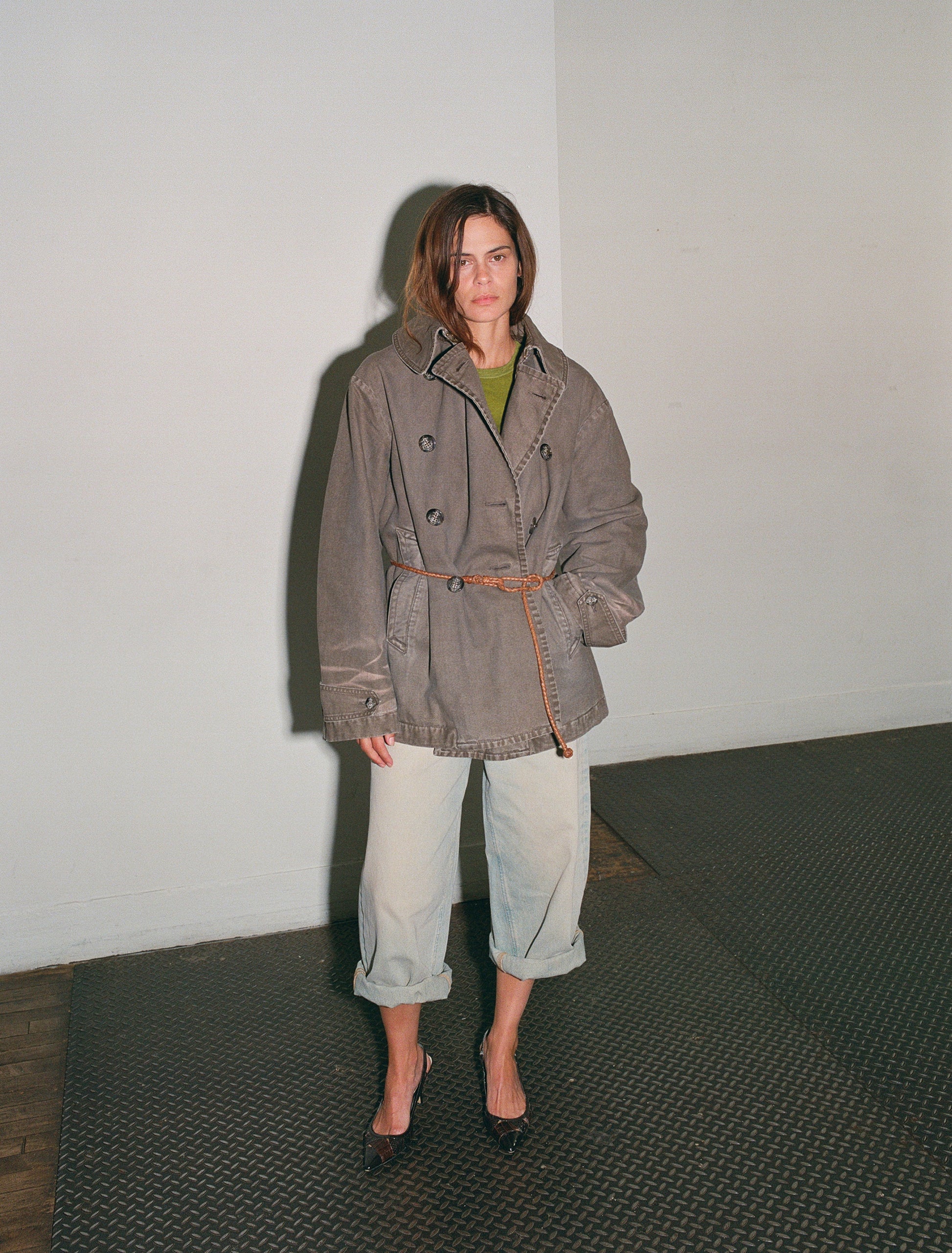 Person wearing a brown coat and light blue jeans standing against a white wall.