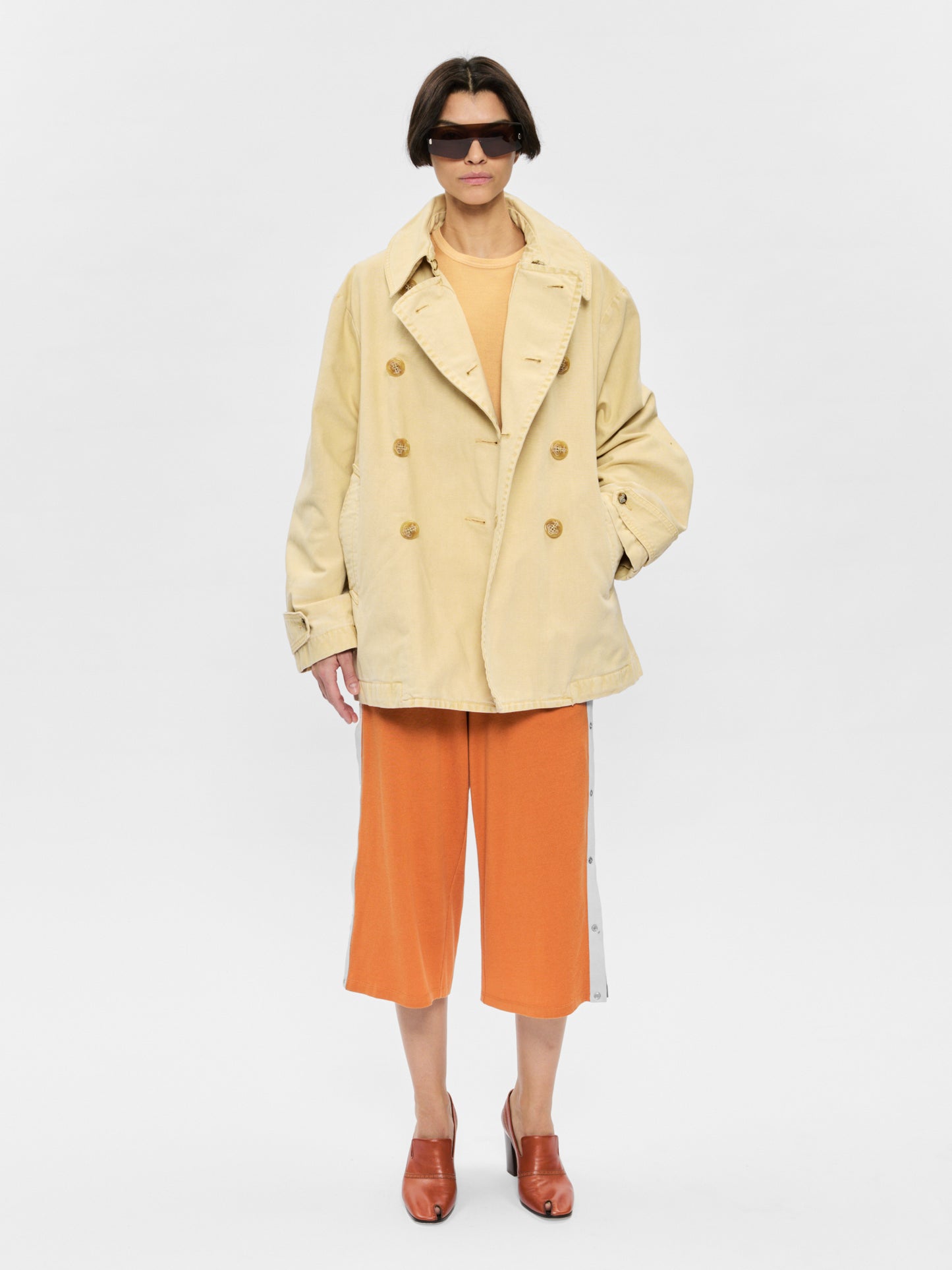 Person wearing a beige trench coat and orange pants on a white background