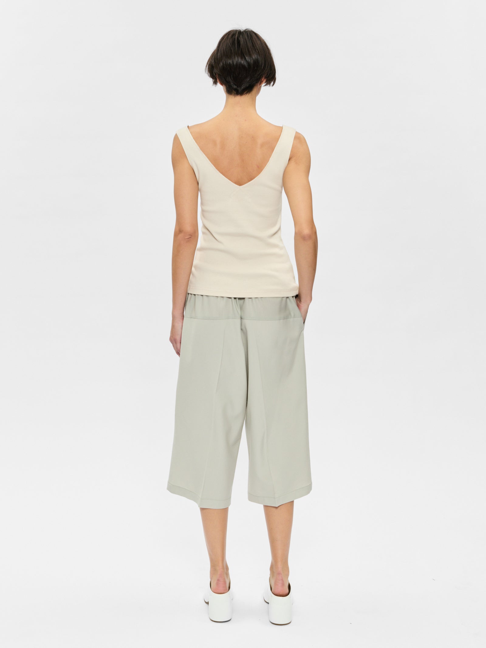 Woman wearing a white sleeveless top and light green pants on a plain background