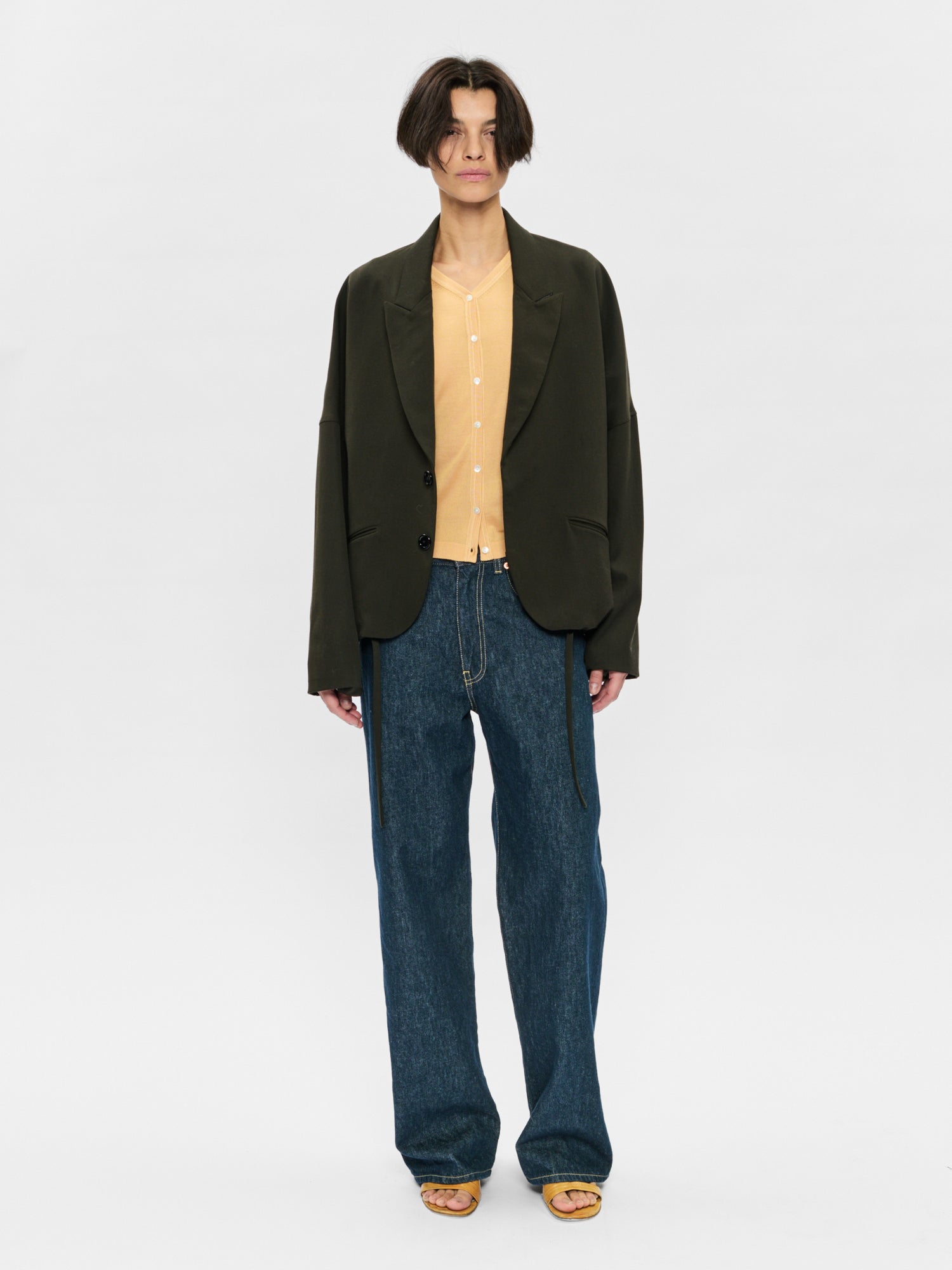 Person wearing a dark green blazer, beige shirt, and blue jeans on a white background