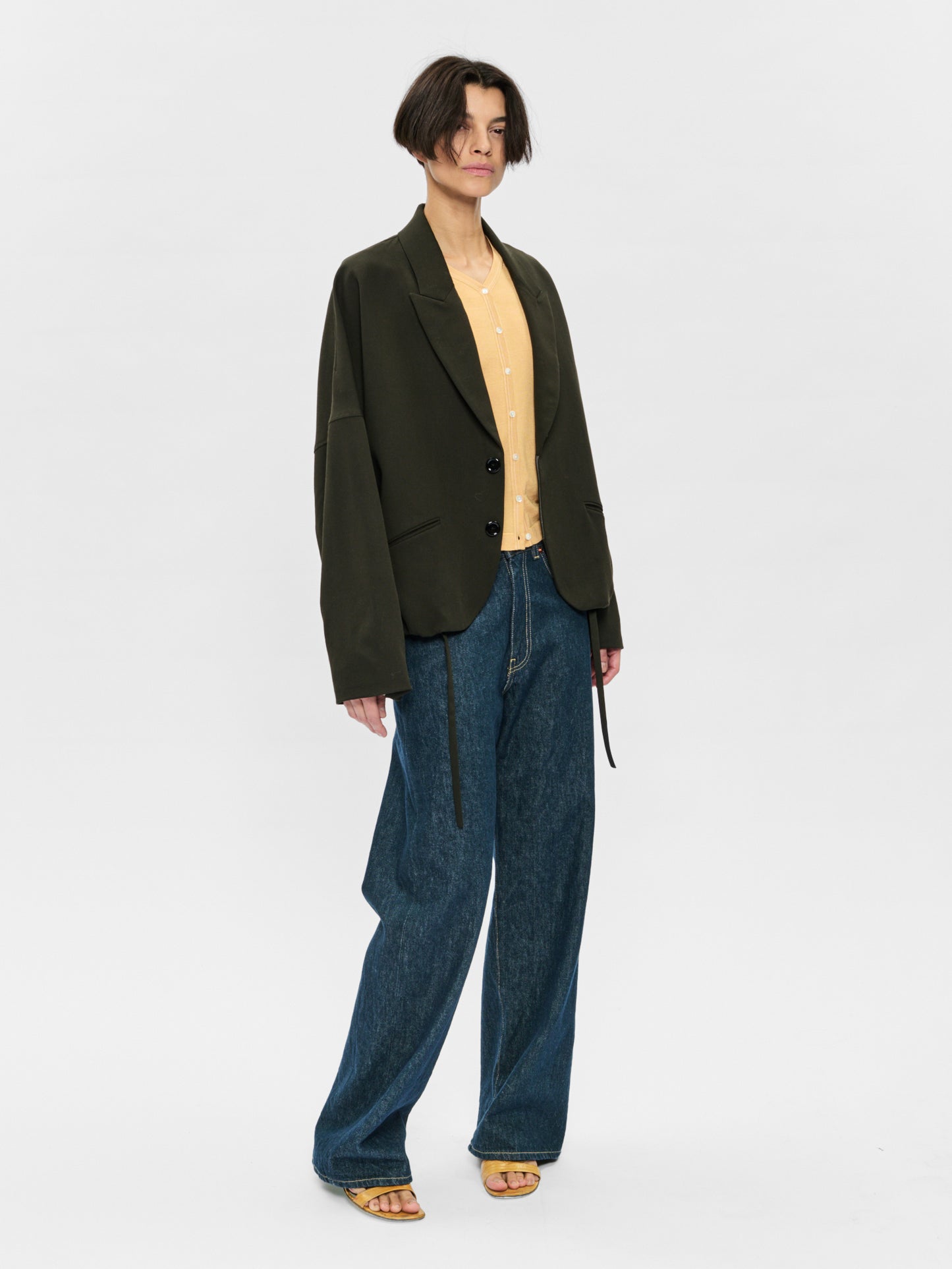 Person wearing a dark green blazer, beige shirt, and blue jeans on a white background