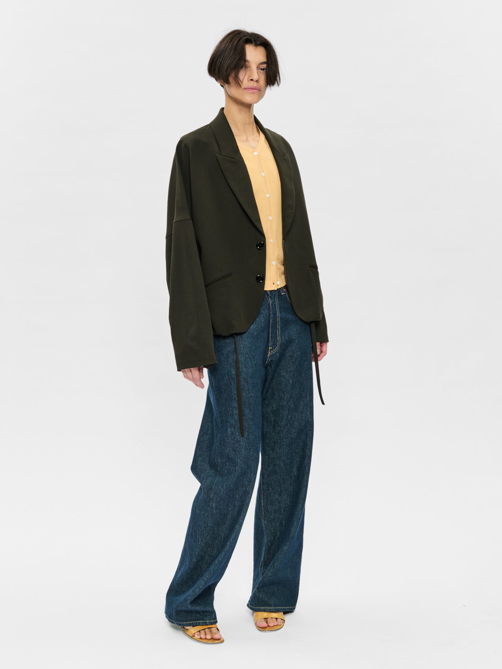 Person wearing a dark green blazer, beige shirt, and blue jeans on a white background