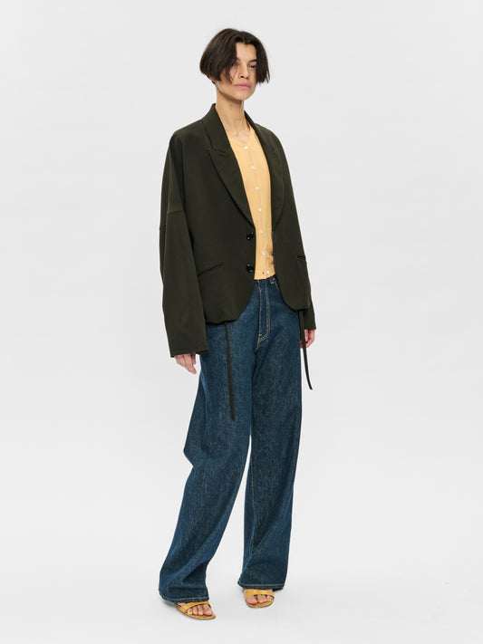 Person wearing a dark green blazer, beige shirt, and blue jeans on a white background